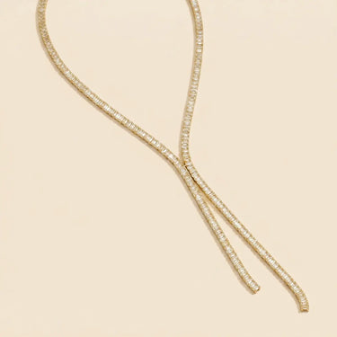 Adele gold baguette tennis necklace with sparkling emerald-cut stones styled on a warm neutral studio background.