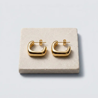 Chunky square gold hoop earrings with a polished mirror finish displayed on a natural stone tray against a soft gray background.