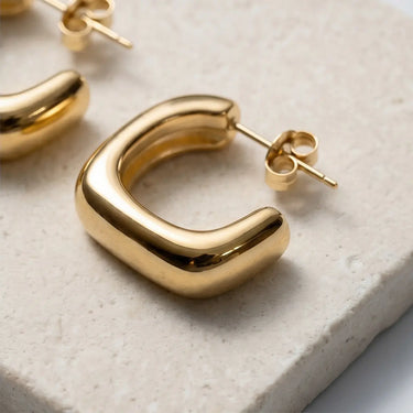 Close-up of chunky gold U-shape hoop earrings with rounded edges and high-shine finish on a textured stone surface.