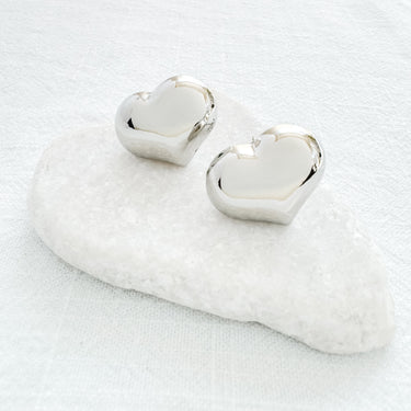 Silver heart-shaped stud earrings displayed on white marble stone, minimalist jewelry photography, Angelina Stud Earrings by Matte.