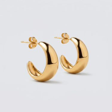 Polished gold crescent hoop earrings with a chunky curved silhouette and mirror-shine finish, photographed on a soft light gray background. Sculptural open hoop design with rounded edges and luxe modern shape.