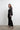 Side view of woman in black satin Astra Crystal Blouse with crystal trim collar and cuffs, styled with black trousers and strappy heels.