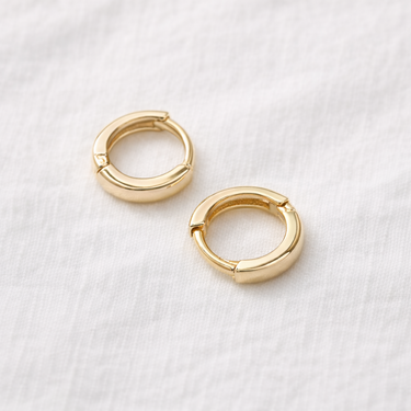 Awen Huggie Earrings – small polished gold huggie hoop earrings with a minimalist design, photographed on a soft neutral background.