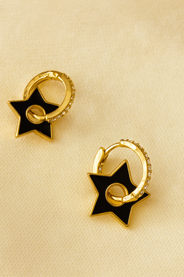 Black Star Huggie Earrings — gold-plated hoops with black enamel star charms, polished mirror finish on beige linen background, Matte Jewelry Collection.