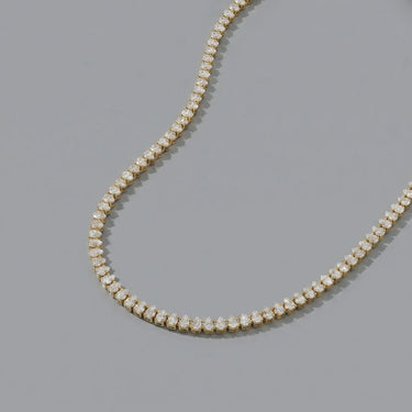 Caelus gold tennis necklace featuring brilliant round-cut crystals in a sleek luxury setting on a soft gray editorial background for an elevated everyday glam look.