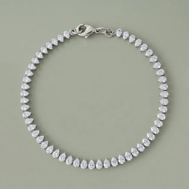 Silver teardrop tennis bracelet with sparkling clear stones and a polished silver clasp on a soft sage gray background, photographed in a clean luxury jewelry editorial style with refined reflections and minimal shadows.