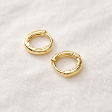 Cassis Huggie Earrings – small polished gold huggie hoop earrings with a smooth minimalist design, photographed on a soft neutral background.