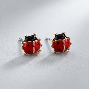 Catarina Stud Earrings featuring glossy red enamel ladybug design with black spots and gold detailing, displayed as a pair on a soft light gray background with subtle shadow, small dainty everyday stud earrings
