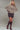 Woman wearing a brown buttoned poncho, patterned shorts, and knee-high boots on a plain background