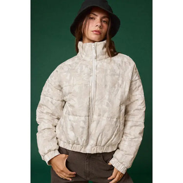 Woman wearing light neutral quilted puff jacket with high neck and elastic cuffs against dark green background.