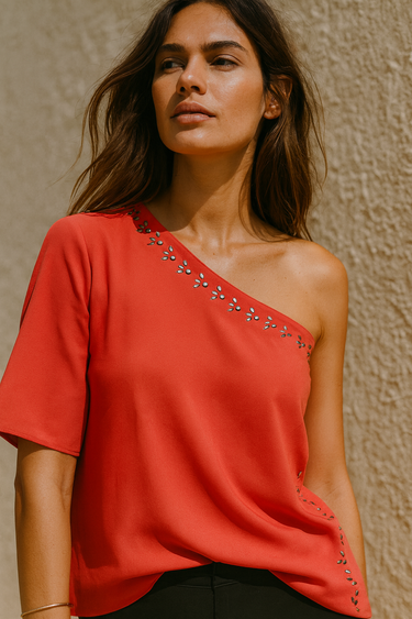 Woman wearing a coral one-shoulder top with embellishments against a beige wall.