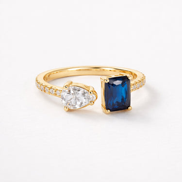 Frills Ring gold open ring with pear cut clear crystal and deep blue emerald cut stone pavé band modern dual stone statement ring on neutral background.