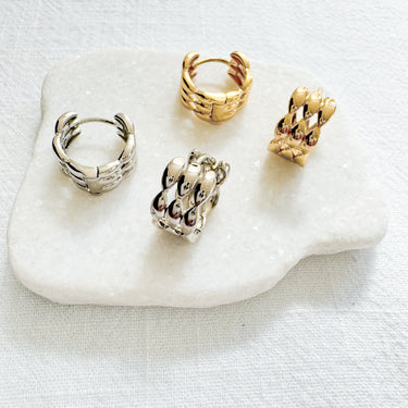 Four Gala Huggie Earrings in gold and silver finishes resting on a white stone against a neutral fabric background.