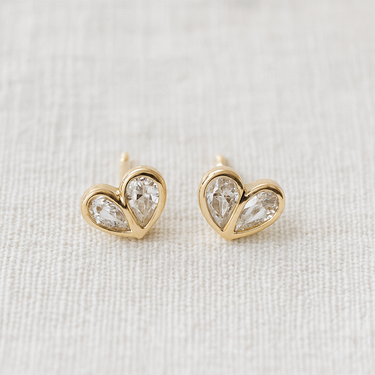 Kuga Stud Earrings featuring gold heart-shaped settings with clear crystals on a soft beige fabric background.