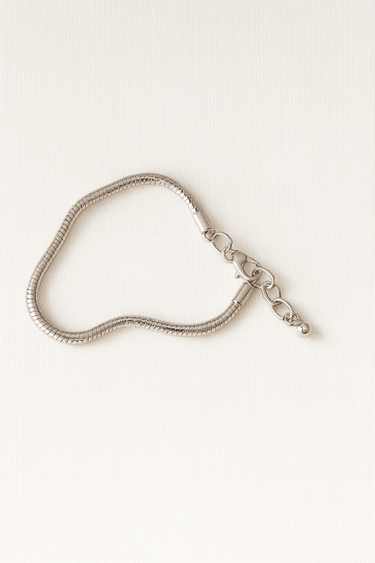 Helios Bracelet — sleek snake-style bracelet in gold or silver with lobster clasp and mirror-polished finish, photographed on beige linen background, Matte Jewelry Collection.