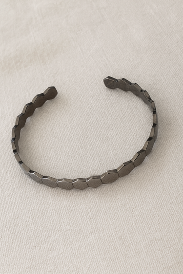 Gunmetal cuff bracelet with geometric hexagonal edges on beige linen background.