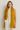 Woman wearing Hono Scarf in mustard yellow knit, styled over white turtleneck and cream pants.