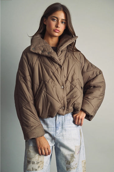 Model wearing the Karls Jacket in taupe, featuring diamond quilting, a padded stand collar, and snap-front closure, styled with light-wash jeans.