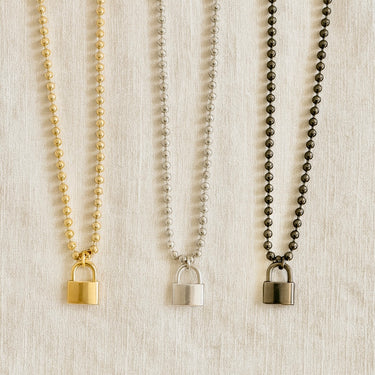 Lock Me Necklaces in gold, silver, and black with padlock pendants displayed on a beige fabric background.