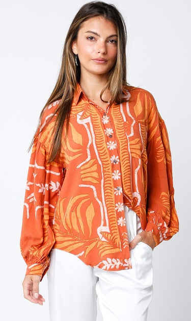 Maréa Blouse sunset-orange statement blouse with artistic tropical-inspired print, button front detail, and relaxed fit long sleeves, styled on model with white pants against clean studio background.
