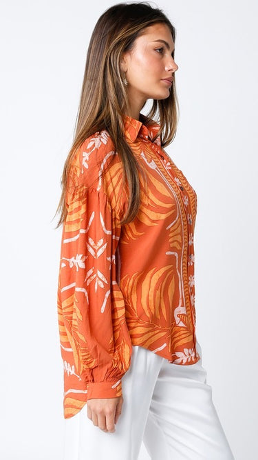 Maréa Blouse side view showing flowy relaxed silhouette, soft volume sleeve shape, and bright orange tropical print detail, styled with white trousers for a vacation-ready look.