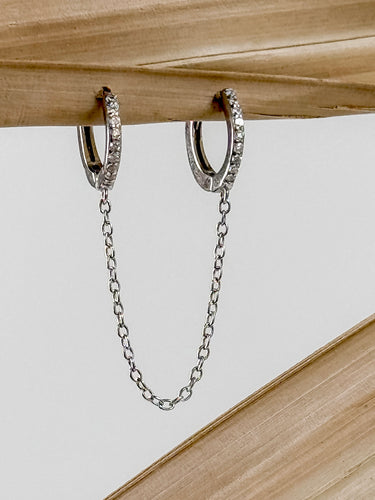 A pair of gold-plated huggie earrings with a chain connecting them, displayed against a wooden background.