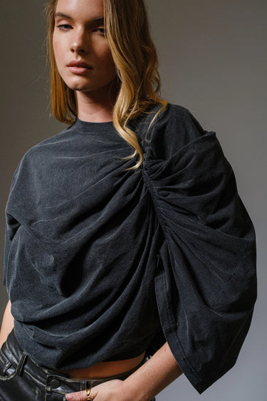 Woman wearing the Roux Top in charcoal black with sculpted ruching and dramatic draped detail, styled in soft natural light on a neutral background.
