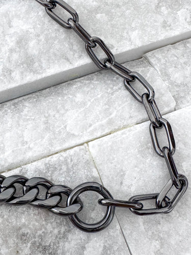 A close-up of a black enamel necklace with a Cuban chain link design placed on a textured surface.