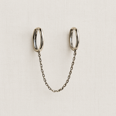 Wagu Huggie Earrings in silver with a connecting chain displayed on a light beige fabric background.