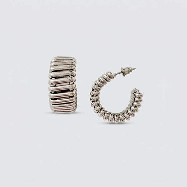 Polished silver ribbed hoop earrings with a chunky sculptural silhouette and high-shine finish on a soft light gray background.