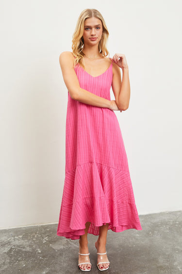 A woman standing in a studio setting, wearing a pink midi dress with spaghetti straps and a ruffled hem.