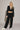 A female model wearing a black Akima pant with a high waist and relaxed fit. The model is paired with a cropped top and white sneakers, standing against a neutral background.