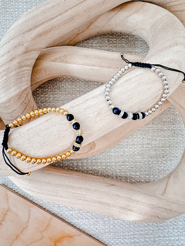 Two bracelets laid on a wooden surface, one with gold-colored beads and the other with black and white beads, showcasing their intricate designs by Matte