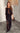 A woman wearing a black maxi dress with sheer details and a beaded design, standing in a room with a minimalist decor.