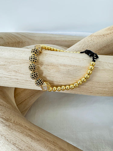 A gold-colored bracelet with a mix of solid and patterned beads displayed on a wooden surface by Matte