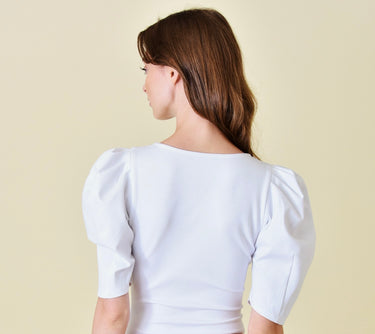 Model wearing the Azura Bodysuit in white — puff sleeve design with deep neckline, styled with camel trousers against a soft beige background, back view.