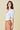 Model wearing the Azura Bodysuit in white — puff sleeve design with deep neckline, styled with camel trousers against a soft beige background, side view.