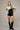 Black fitted bodysuit with metallic contour embroidery and deep V neckline, styled with a draped jacket on a model in an editorial neutral setting.