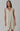 A sleeveless, beige linen dress with fringed detailing at the V-neckline and a raw hem, worn by a model.