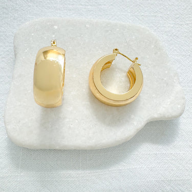 Ixelles Huggie Earrings in polished gold finish resting on a white stone against neutral fabric background.