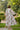 A maxi dress with a floral pattern in pink and green on a white background, worn by a model facing away from the camera, standing in a garden setting.