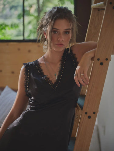 A woman standing next to a ladder, wearing a black satin sleeveless dress with a V-neckline and lace trim, paired with black boots.