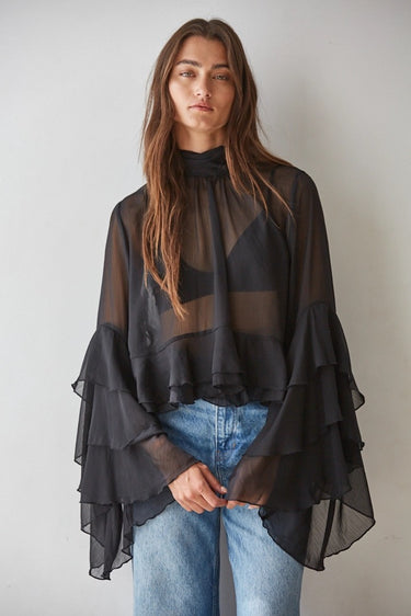 Woman wearing the Maruk Blouse in black, featuring a lightweight sheer fabric, relaxed silhouette, elegant high neckline, dramatic tiered bell sleeves, and flowing design styled with blue denim jeans.