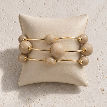 Minutti Bracelet in neutral beige featuring polished round beads and slim gold-tone bars in a sculptural multi-row design displayed on a cream jewelry cushion.