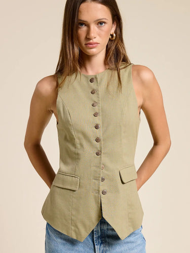 Model wearing the Muse Vest in soft sage green featuring a tailored fitted silhouette, button-front closure, pointed hem, sleeveless cut, and decorative flap pockets paired with light wash denim jeans.