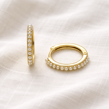Naia Huggie Earrings – slim gold huggie earrings with pearl detailing, photographed on a soft neutral fabric background.