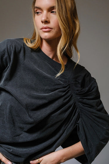 Woman wearing the Roux Top in charcoal black with sculpted ruching and dramatic draped detail, styled in soft natural light on a neutral background.