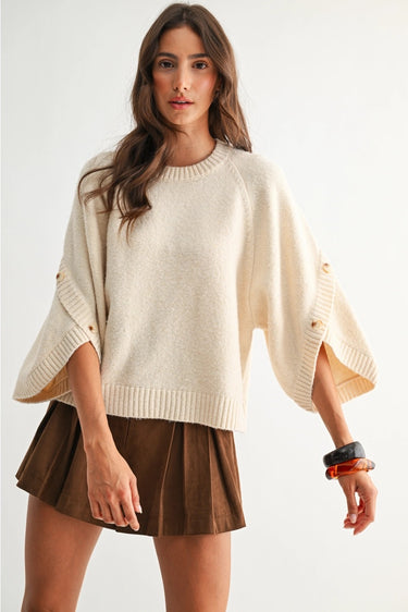 Sienu Sweater in cream featuring a relaxed oversized knit silhouette with cape sleeves, decorative button details, and a soft draped fit paired with a brown mini skirt.