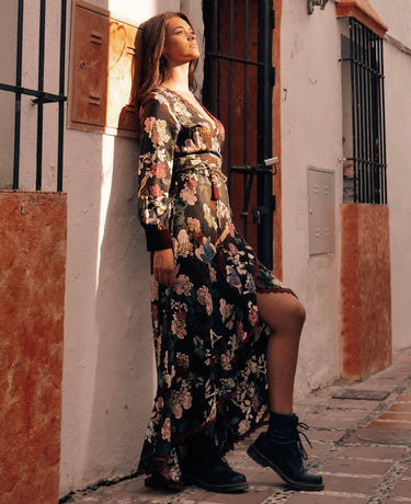A person wearing a floral maxi dress with long sleeves, styled with a belt, and black boots, standing in a narrow alley with buildings in the background.