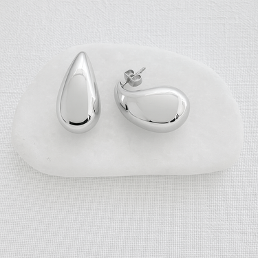 Polished silver teardrop Trena stud earrings resting on a white stone against a neutral fabric background.